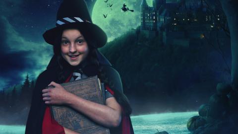 A girl wearing a witches hat and cloak stands in a misty forest with a mountain and castle in the background under a full moon-lit sky.