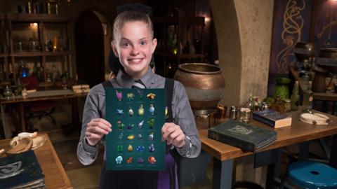 A girl with blonde hair in a potions classroom is holding a sheet of paper with potions ingredients on it, Ethel from The Worst Witch.