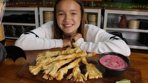 A girl sitting next to a kitchen counter with cheesy pastry strips in the shape of witches broomsticks.