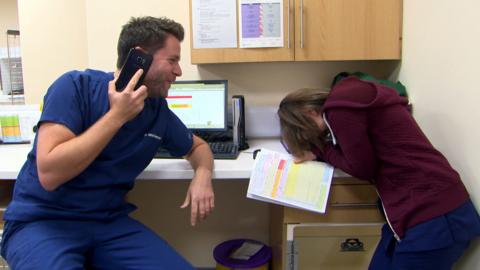 A man dressed in dark blue scrubs laughs while talking on the phone, the woman sitting next to him is doubled up with laughter.