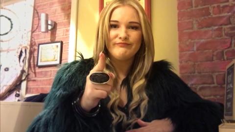 A girl with long blonde hair and a furry jacket points at the camera and smiles.