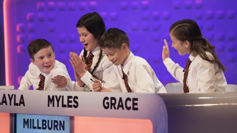 Four school children sat behind a quiz show desk, (Millburn School team).