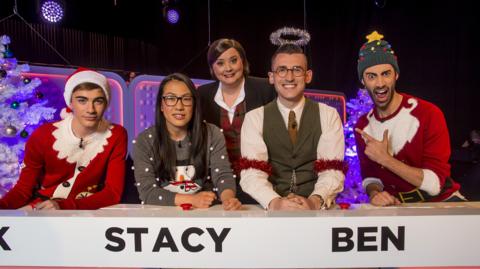 Five people stand behind a quiz show desk, Susan Calman with Team Sprouts made up of Nick James, Stacy Liu, Ben Shires and Richard Caine.