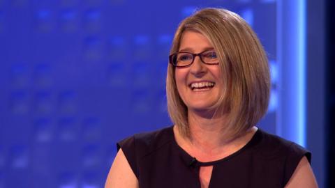 A blonde female teacher wearing a black dress and glasses smiling in front of a blue background.