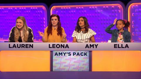 Three women and a girl sit behind a gameshow studio desk.