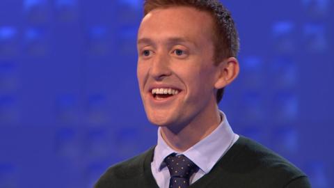 A teacher wearing a green jumper and a tie laughs.