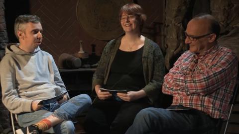 A woman and two men sat in a low lit room next to each other.