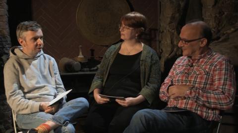 A woman and two men sat in a low lit room next to each other.