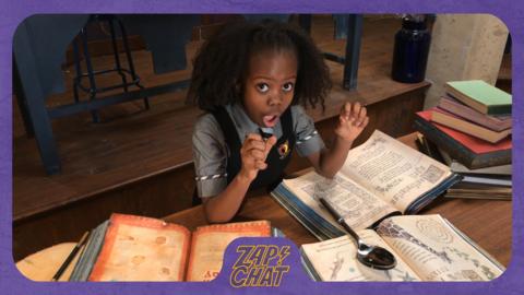 A girl at a desk with open books, looking shocked. Beatrice from The Worst Witch.