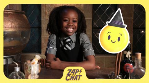 A girl sat at a desk looking happy. A smiley emoji is next to her. Beatrice from The Worst Witch.