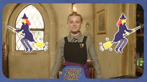 A young witch in Cackle's Academy uniform (Sybil from The Worst Witch) smiles with two witches on brooms as emojis flying on either side of her.