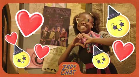 A young witch (Bea) makes a heart shape using her hands, wearing sunglasses, surrounded by heart emojis.