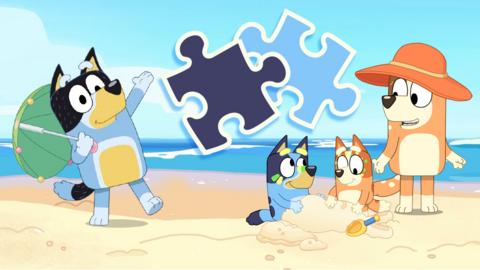 Bandit is holding a sunshade, whilst Mum wears a sun hat and Bluey and Bingo are playing in the sand on the beach. There are two jigsaw piece shapes in the image.