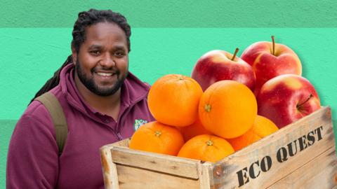 Ranger Hamza is smiling next to a wooden crate of different fruits