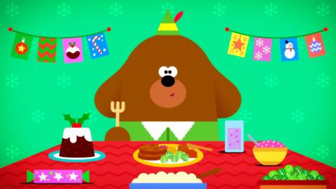 Duggee is sitting at a dinner table covered in festive food. Duggee is wearing an elf costume.