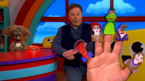 Justin Fletcher is sat opposite of Dodge from CBeebies House are both looking to camera. A large hand is beside them with Justin's House characters on each finger and thumb