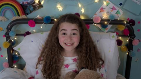 Molly is sitting on her bed, looking up at the camera and smiling.