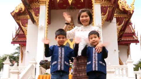 Two boys are with their mother, waving to the camera.