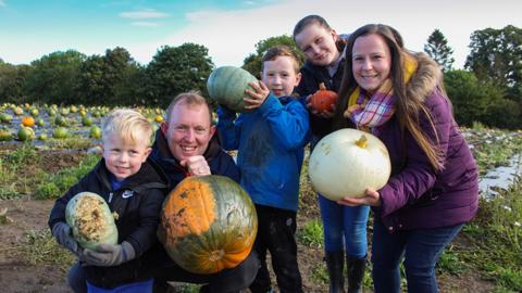 A family of five holding up various sized pumpkins in a field.