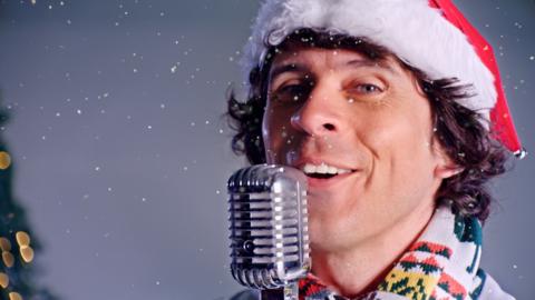 Close up of Andys face, smiling, next to a microphone. He is wearing a red and white santa hat.