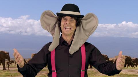 Andy wearing a hat and big elephant ears in front of a wildlife scene of elephants.