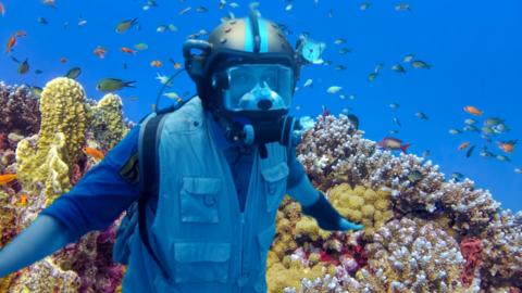 Andy wearing his scuba diving helmet in front of the coral reef surrounded by fish.