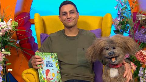 Ben and Dodge are sat on a yellow chair in CBeebies house, ben is holding a book and both are smiling to camera
