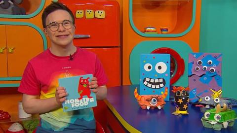 George is sat in the CBeebies House kitchen holding a book. Beside him is a table with homemade monsters on it.