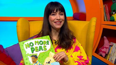 Rebecca is sat on a yellow chair in CBeebies house, Rebecca is holding a book that says 'No More Peas' and is smiling to camera