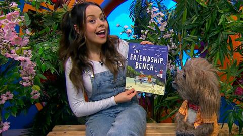 Evie and Dodge are sat on a bench in the CBeebies house. Evie is holding a book called 'The Friendship Bench' and is smiling to camera. Dodge is looking excitedly at the book Evie is holding.