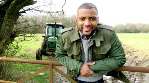 JB from Down On The Farm in a field leaning on a gate in front of a tractor.