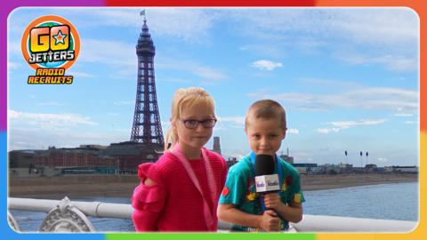 Go Jetters Radio Recruits Clarissa and Luis  are standing on a pier in Blackpool, with the beach and Blackpool Tower in the background.