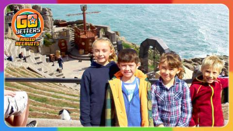 Go Jetters Radio Recruits Niall, Issy, Henry and Morgan are standing in the seating area of the Minack Theatre, with the sea and stage in the background.