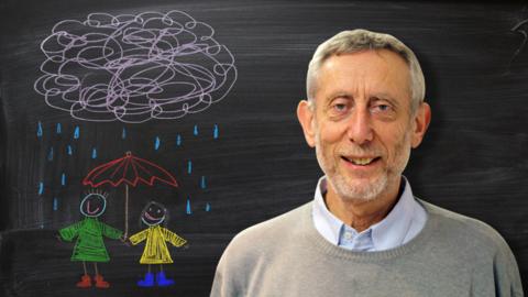 Michael Rosen from Poetry Playtime in front of a blackboard picture of people carrying an umbrella under a raincloud.