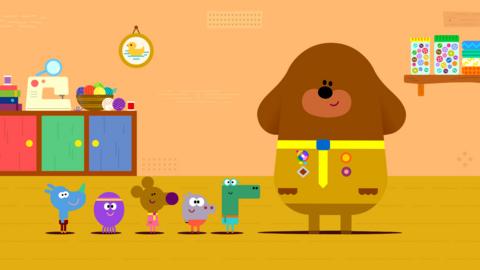 Duggee and the Squirrels are standing in a room. Duggee is wearing his regular yellow outfit and in the background are many crafting items like buttons, a sewing machine, and wool.