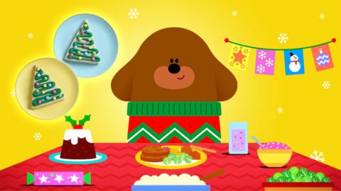 Duggee in his Christmas jumper at the dinner table with lots of delicious snacks especially the tree cookies he baked.