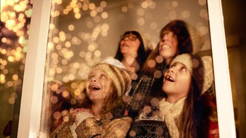 Two young girls and their mothers are looking outside a window in amazement at fireworks in the sky.