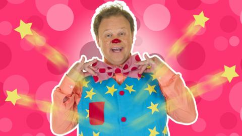 Mr Tumble is holding his bowtie and smiling at the camera as four stars are shotting off his waistcoat.