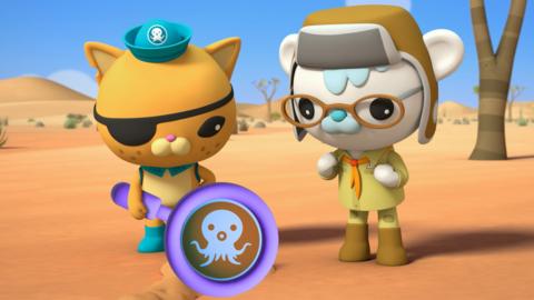 Kwazii and Tracker from Octonauts: Above & Beyond are stood next to one another in an orange desert. They are both looking down at the sand, Kwazii has a large purple magnifying glass in hand highlighting the Octonauts logo.