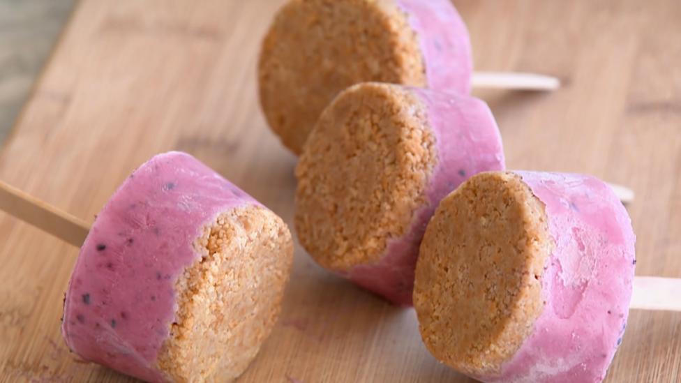 Four bright pink yoghurt lollies with a biscuit base resting on a wooden board.