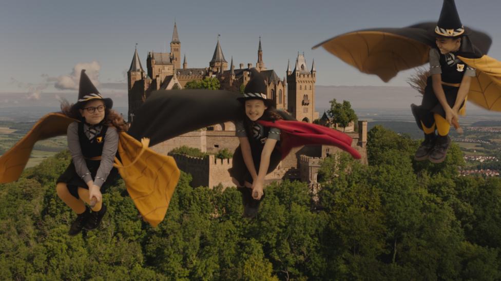 Three witches fly in the air on broomsticks in front of a castle.