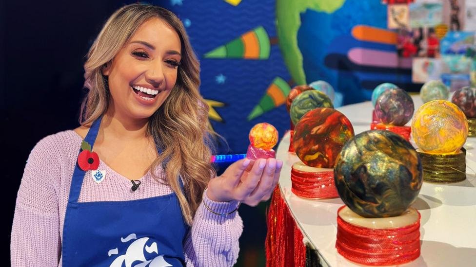 Nikita Kanda, a woman of Indian descent with blonde hair is smiling brightly holding up a small painted tealight in the Blue Peter studio.