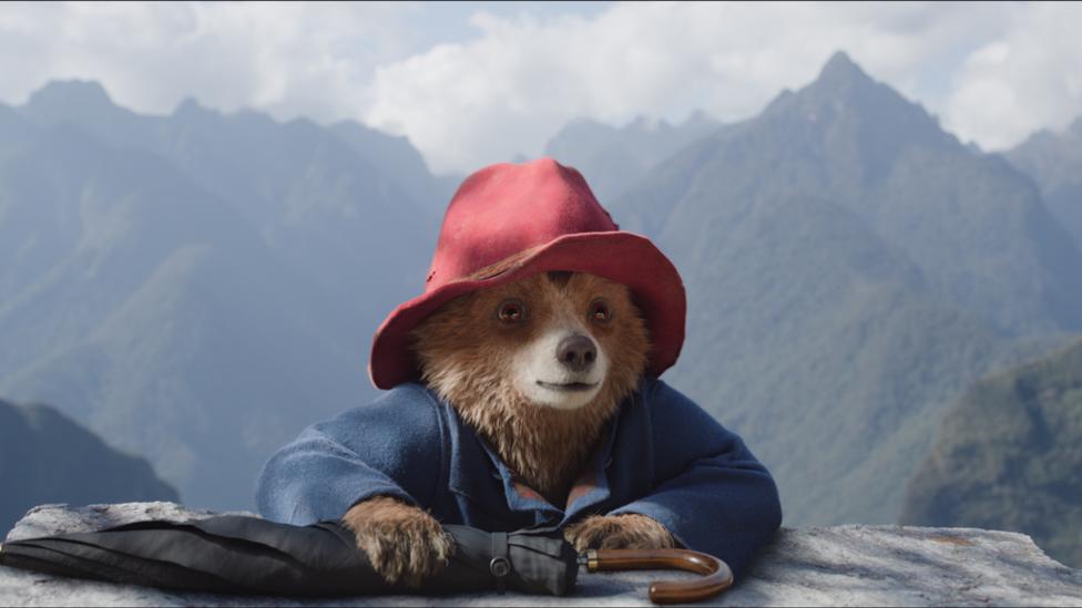 Paddington bear climbing a mountain holding his umbrella and wearing his signature red hat.