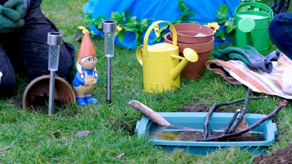 Image of a completed toad abode featuring a bucket of water with sticks and stones, a plant pot house and a garden gnome.