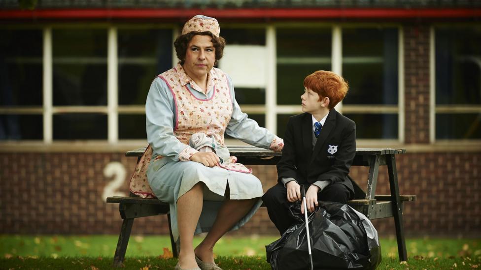 A ginger school boy is sitting next to an old lunch lady on a park bench