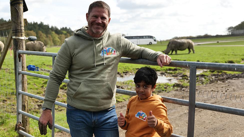 An image of Steve stood next to Aneeshwar, behind them is a rhino enclosure. Steve has mud on his hand.