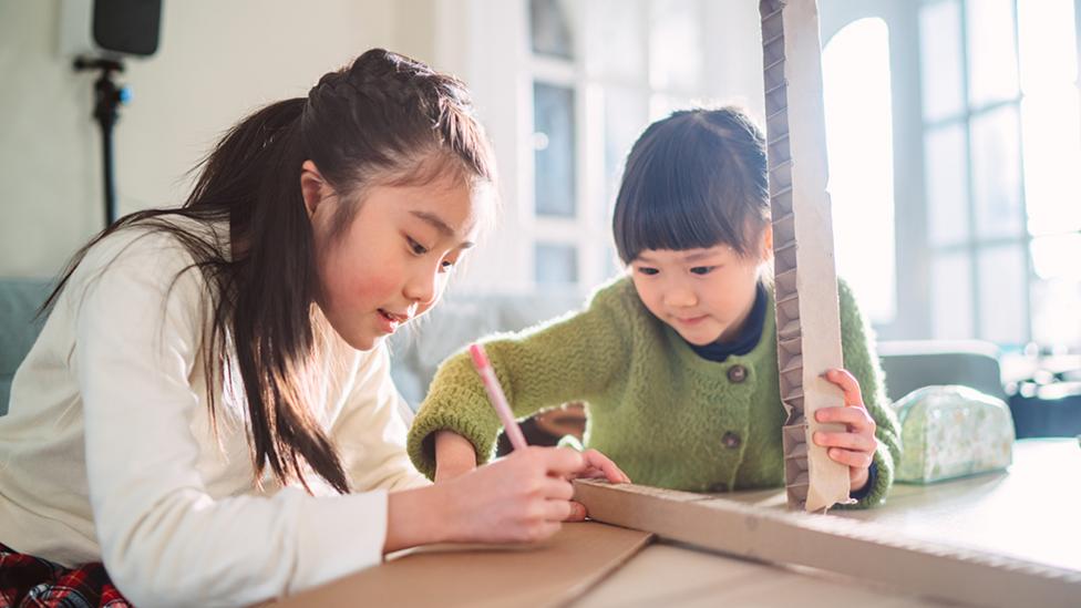 Two young sisters are sitting at a desk creating cardboard crafts.