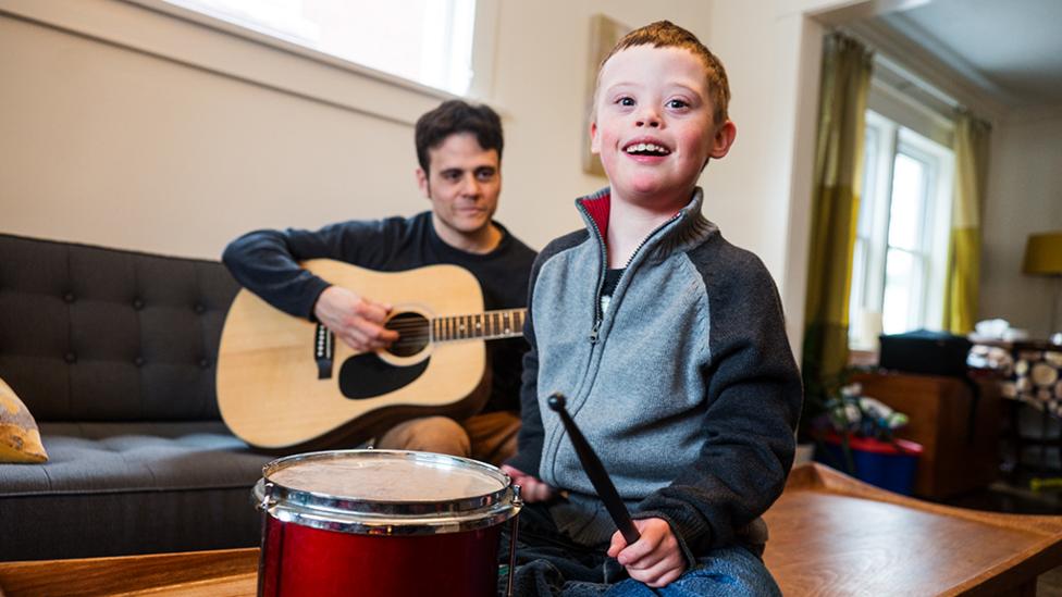 A young boy is smiling holding a drum, with an adult sitting in the background holding a guitar.