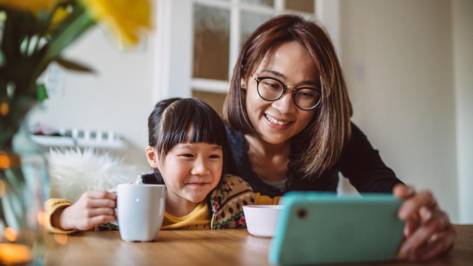 A mother and young daughter are sitting down at a table, looking at a mobile phone.