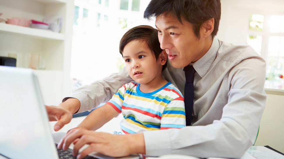 A young boy sat on his father's lap, at a table. The father is typing and showing the little boy the laptop screen.